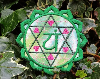 May include: Embroidered patch featuring a green and pink design with a central geometric pattern and a stylized symbol. The patch has a scalloped edge and is set against a backdrop of green foliage.