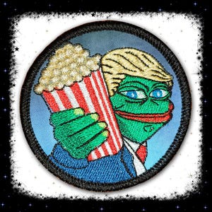 May include: An embroidered patch depicting a green cartoon frog wearing a blue suit and a red tie. The frog is holding a red and white striped bucket of popcorn. The frog has a surprised expression on its face.