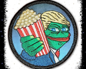 May include: An embroidered patch featuring a green cartoon frog wearing a blue suit and a red tie. The frog is holding a red and white striped bucket of popcorn. The frog has a surprised expression on its face.