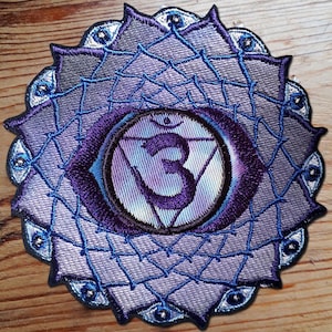 May include: Purple and blue embroidered patch featuring a stylised eye symbol in the centre. The symbol is surrounded by a circle of smaller eyes. The patch is designed to represent the sixth chakra, the third eye chakra.