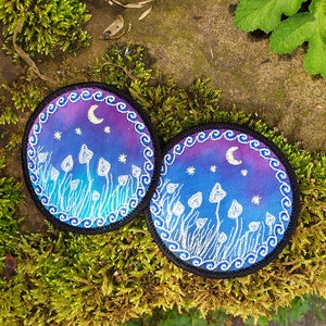 May include: Two round embroidered patches with a night sky scene. The patches feature a crescent moon, stars, and white mushrooms on a purple and blue gradient background. The patches have a black border with a white wavy design.