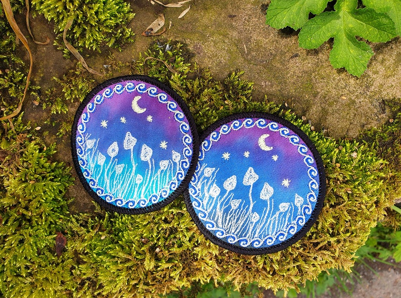 May include: Two round embroidered patches with a night sky scene. The patches feature a crescent moon, stars, and white mushrooms on a purple and blue gradient background. The patches have a black border with a white wavy design.