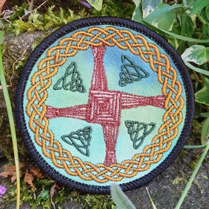 May include: A circular embroidered patch featuring a central red St. Brigid's cross on a light blue background. The design is surrounded by a golden yellow Celtic knot border and accented with four small dark green triquetra symbols.