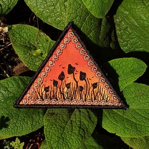 May include: A triangular patch with a black outline and a gold glitter border. The patch features a sunset scene with a field of black silhouette mushrooms.