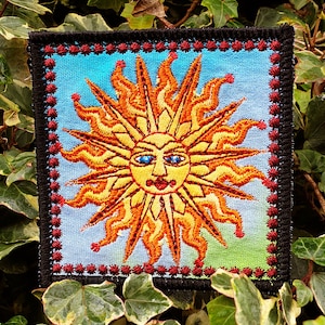 May include: A colorful embroidered patch featuring a stylized sun with a smiling face and blue eyes. The sun is surrounded by a red and black border.