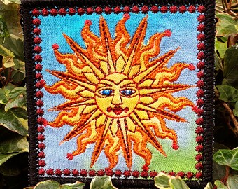 May include: A colorful embroidered patch featuring a stylized sun with a smiling face and blue eyes. The sun is surrounded by a red and black border.