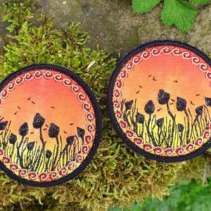May include: Two round fabric coasters with a black border and a gold and red embroidered design. The center of each coaster features a sunset scene with a field of tall, black silhouettes of mushrooms.