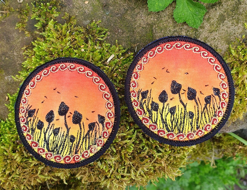 May include: Two round fabric coasters with a black border and a gold and red embroidered design. The center of each coaster features a sunset scene with a field of tall, black silhouettes of mushrooms.