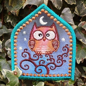 May include: Embroidered patch featuring a brown owl with large eyes, perched on a brown tree branch with swirling vines. The owl is set against a blue background with a crescent moon and stars. The patch is surrounded by a turquoise border with gold accents.