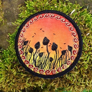 May include: A round embroidered patch with a sunset scene and a field of black mushrooms. The patch has a gold and red border with a swirling pattern.