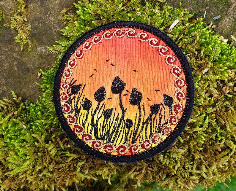 May include: A round embroidered patch with a sunset scene and a field of black mushrooms. The patch has a gold and red border with a swirling pattern.