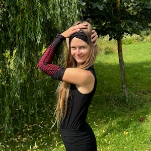 May include: A woman wearing a black sleeveless top with a black headband and red and black arm warmers. She is standing in a grassy area with trees in the background.