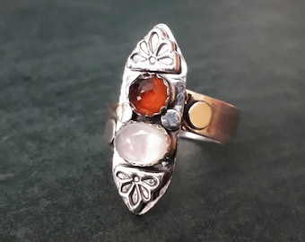 May include: A silver ring with a long, pointed, oval-shaped setting. The setting is decorated with floral designs and features three stones: a large, round, white stone, a smaller, round, orange stone, and a tiny, round, clear stone.