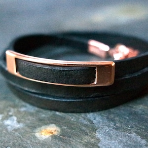 Pode incluir: Um bracelete de couro preto com um detalhe de barra em ouro rosa. O bracelete é feito de três camadas de couro e tem um design simples e moderno.