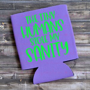 May include: A purple can cooler with the text "THE TINY HUMANS STOLE MY SANITY" in bright green. The cooler is made of a foam-like material and has a cutout for a can or bottle. The background is a wooden surface.