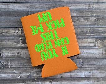 May include: Orange can cooler with the text "IF YOU CAN READ THIS PICK ME UP!" in bright green. The cooler is made of a textured material and is designed to keep beverages cold. The background is a rustic wooden surface.