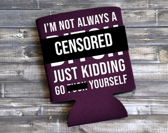 May include: A dark purple can cooler with white text that reads "I'M NOT ALWAYS A CENSORED JUST KIDDING GO CENSORED YOURSELF". The text is partially obscured by a black bar. The cooler is on a wooden surface.