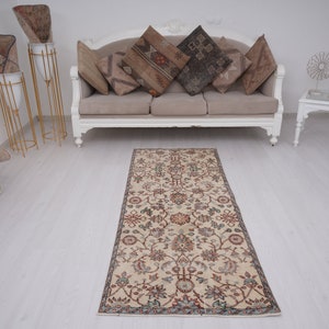 May include: A cream-colored runner rug with a floral pattern in shades of red, green, and blue. The rug is laid out on a white wood floor in front of a white sofa with pillows.