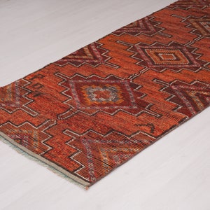 May include: A long, narrow rug with a geometric pattern in shades of red, orange, and brown. The rug has a fringe border.