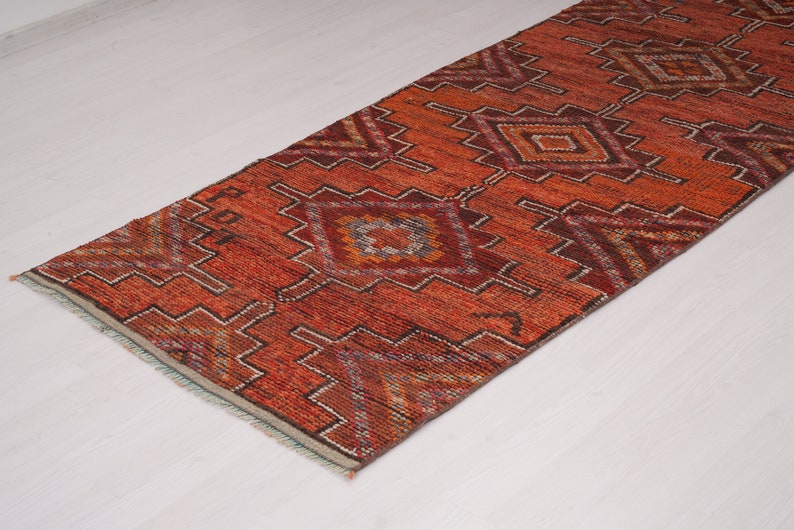 May include: A long, narrow rug with a geometric pattern in shades of red, orange, and brown. The rug has a fringe border.