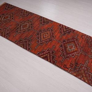 May include: A long, narrow rug with a geometric pattern in shades of red, orange, brown, and white. The rug has a fringed edge.
