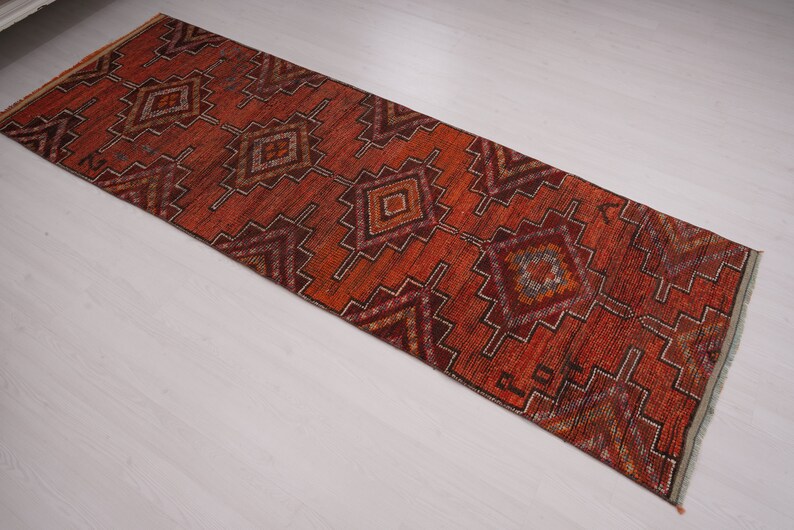 May include: A long, narrow rug with a geometric pattern in shades of red, orange, brown, and white. The rug has a fringed edge.