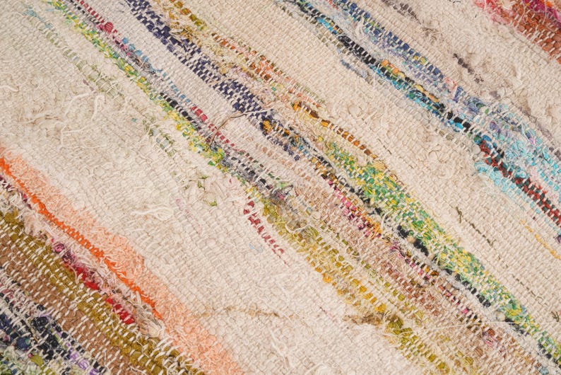 May include: A woven rug featuring a textured, off-white base with diagonal stripes of multicolored fabric strips. The textile displays a variety of vibrant hues including orange, blue, green, yellow, and red, integrated into the weave.