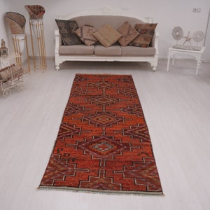 May include: A red, orange, and brown patterned runner rug with a geometric design. The rug is laid out on a white wood floor in a room with white furniture.