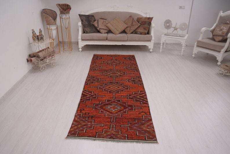 May include: A red, orange, and brown patterned runner rug with a geometric design. The rug is laid out on a white wood floor in a room with white furniture.