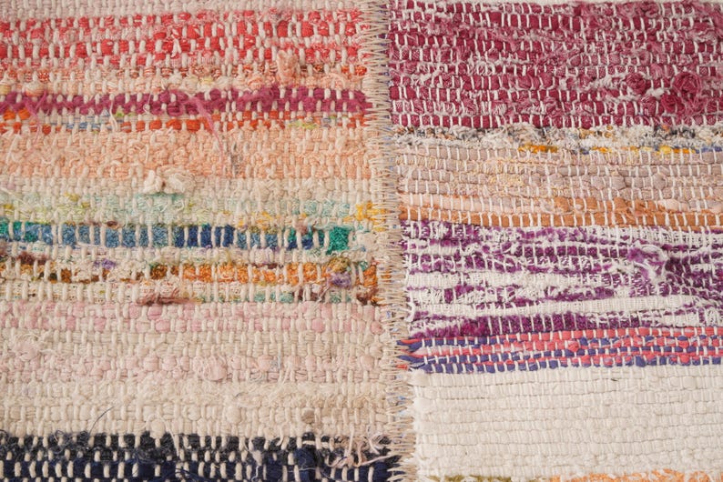 May include: A woven rag rug featuring a patchwork design with two distinct sections joined by a vertical seam. The fabric strips display a multicolored palette including shades of pink, orange, blue, white, and purple in a textured weave.