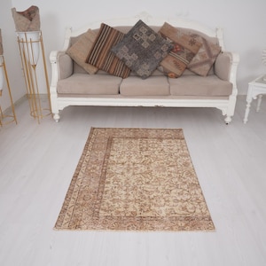 May include: A rectangular, beige and brown patterned rug with a detailed floral design. The rug is placed on a white floor in front of a beige sofa with decorative cushions. The room has a neutral colour palette.