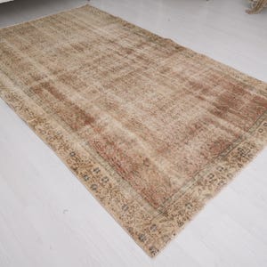 May include: A rectangular, distressed-look rug with a faded, vintage appearance. The rug features a central field of muted brown and beige tones, with a reddish-brown border and subtle floral patterns. The rug is placed on a white surface.