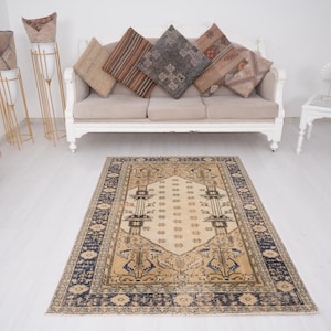 May include: A beige and blue patterned rug with a cream-colored center. The rug is laid out on a white floor in front of a white couch with beige cushions.