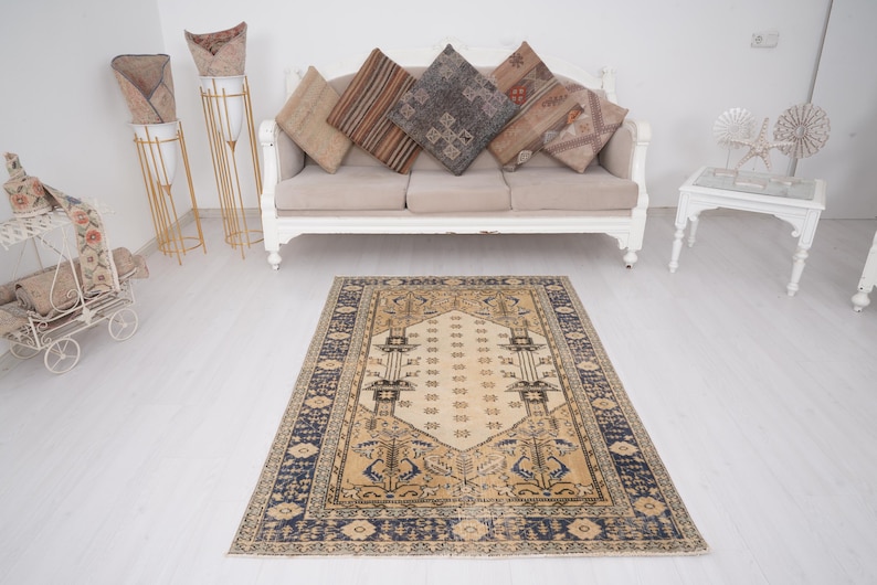 May include: A beige and blue patterned rug with a cream-colored center. The rug is laid out on a white floor in front of a white couch with beige cushions.