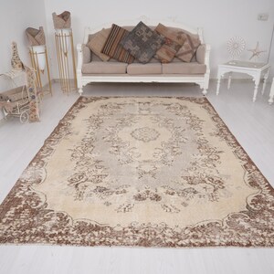 May include: A large, rectangular rug with an ornate floral pattern in beige, brown, and cream tones. The rug features a central medallion design and a detailed border. It is placed on a light-coloured wooden floor, with furniture in the background.