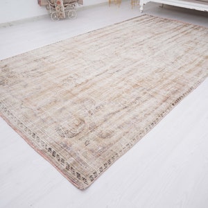 Op de afbeelding: Een licht beige vloerkleed met een vervaagd bloemenpatroon en een roze rand. Het kleed heeft een verweerde, vintage uitstraling.