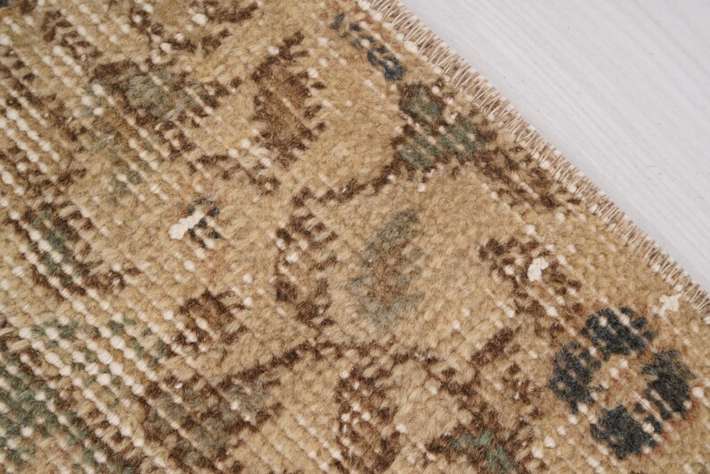 May include: Close-up of a section of a beige and brown rug with a distressed, vintage appearance. The rug features a repeating geometric pattern with accents of green and blue. The edges of the rug are visible, showing a fringed border.
