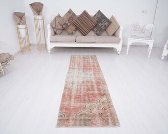 May include: A long, rectangular, vintage-style rug with a faded pink and cream color scheme. The rug has a distressed, worn look with intricate patterns throughout. It is laid out on a white floor in front of a white sofa.