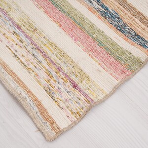 May include: A rectangular woven rug featuring a series of vertical stripes in various colors including pink, green, blue, yellow, and orange, interspersed with wide bands of off-white fabric, creating a textured and multi-colored striped pattern.
