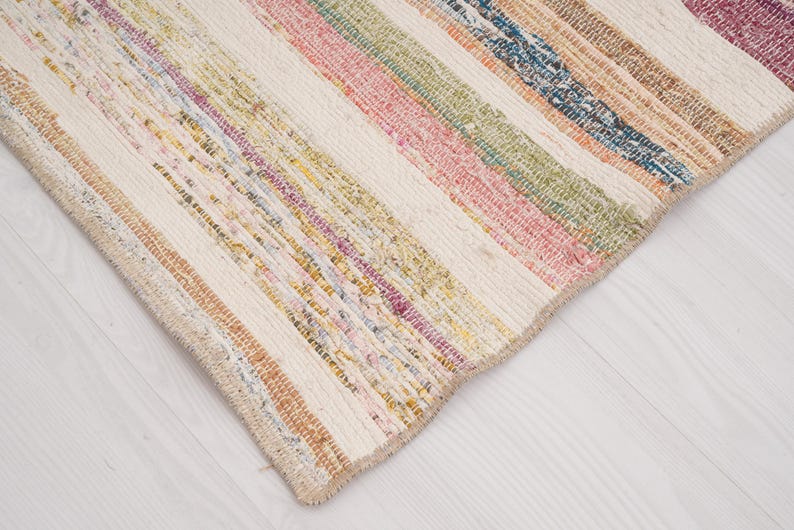 May include: A rectangular woven rug featuring a series of vertical stripes in various colors including pink, green, blue, yellow, and orange, interspersed with wide bands of off-white fabric, creating a textured and multi-colored striped pattern.
