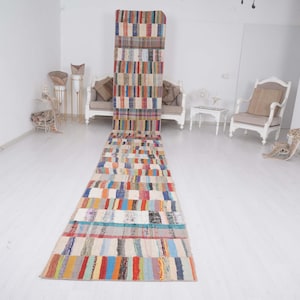 Pode incluir: Um tapete passadeira longo e retangular com um padrão de riscas multicoloridas, composto por blocos de tiras de tecido vermelhas, azuis, amarelas, verdes e brancas. O tapete está posicionado verticalmente, estendendo-se do chão até um sofá.
