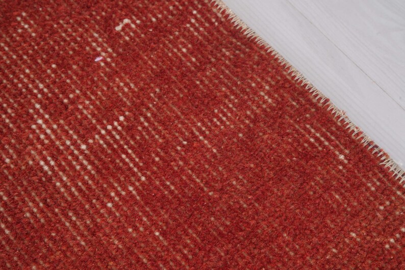 Puede incluir: Una alfombra de color rojo intenso con textura que presenta una superficie de pelo denso y anudado con sutiles variaciones verticales en la intensidad del color y un borde cosido reforzado de color claro a lo largo del borde diagonal.