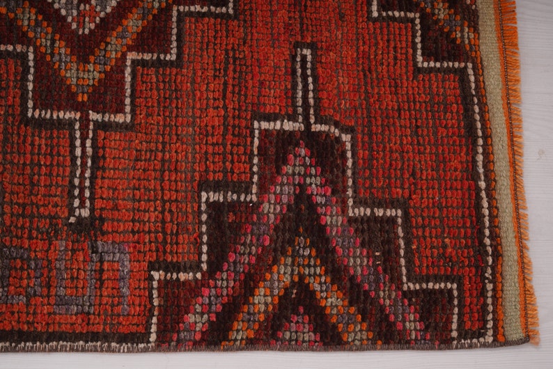 May include: Close-up of a red, orange, and brown patterned rug with a fringe border. The rug has a geometric design with a dark brown center.
