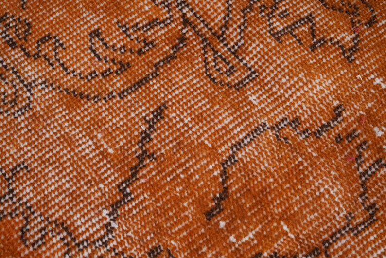 Puede incluir: Un tejido con un color base naranja quemado vibrante que presenta un patr&oacute;n repetitivo de motivos lineales oscuros y dentados en marr&oacute;n, junto con peque&ntilde;as motas blancas distribuidas por toda la superficie, creando un aspecto texturizado.