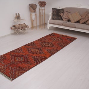 May include: A red, orange, and blue patterned runner rug with a geometric design. The rug is laid out on a white floor in a room with white walls.
