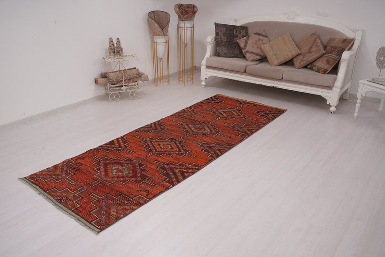 May include: A red, orange, and blue patterned runner rug with a geometric design. The rug is laid out on a white floor in a room with white walls.