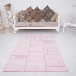 May include: A pink patchwork rug with a distressed look, featuring a variety of rectangular shapes and textures. The rug is laid out on a white wooden floor in a living room setting.