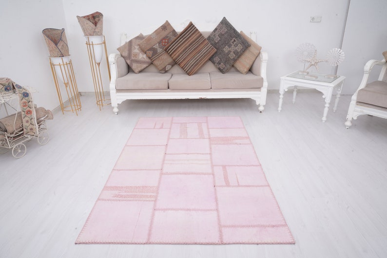 May include: A pink patchwork rug with a distressed look, featuring a variety of rectangular shapes and textures. The rug is laid out on a white wooden floor in a living room setting.