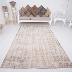 Op de afbeelding: Een lichtbeige vintage tapijt met een verweerde look. Het tapijt heeft een subtiel patroon en een verbleekte uitstraling, wat het een rustieke charme geeft. Het ligt op een witte vloer in een kamer met witte meubels.