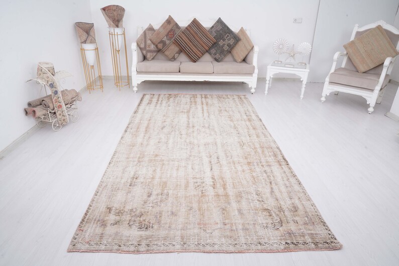 Op de afbeelding: Een lichtbeige vintage tapijt met een verweerde look. Het tapijt heeft een subtiel patroon en een verbleekte uitstraling, wat het een rustieke charme geeft. Het ligt op een witte vloer in een kamer met witte meubels.
